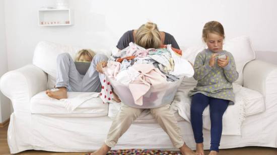 Exhausted mother with laundry basket on couch with children using digital tablet and cell phone