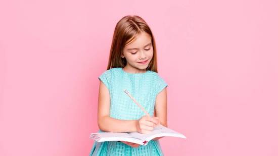Lovely adorable charming little girl is looking down at the copybook in her hands and writing information there, she is wearing light blue dress, isolated on bright pink background, copyspace
